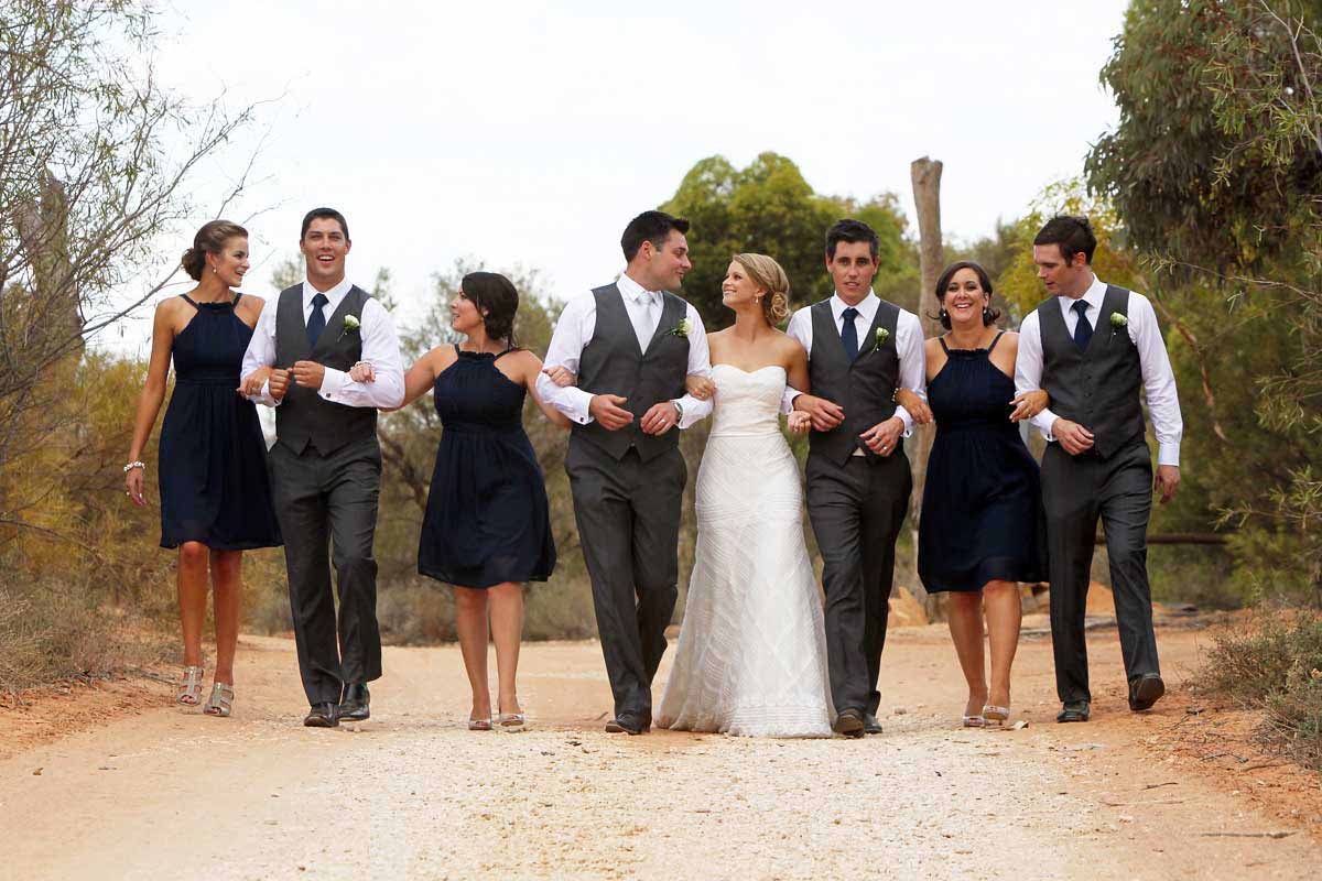 Wedding party of bride in white dress and bridal party in navy blue walking arm-in-arm along dirt path through native Australian bushland at Pike River Villas