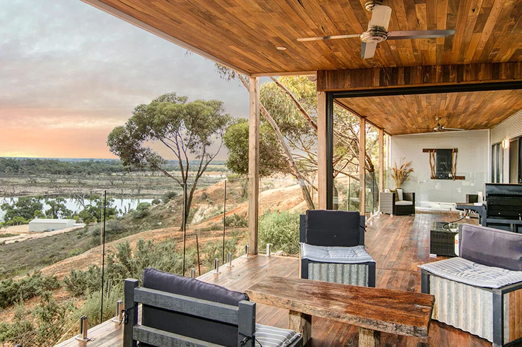 Covered outdoor dining deck with timber ceiling, glass balustrade overlooking Murray River at sunset, featuring modern furniture and indoor-outdoor living space