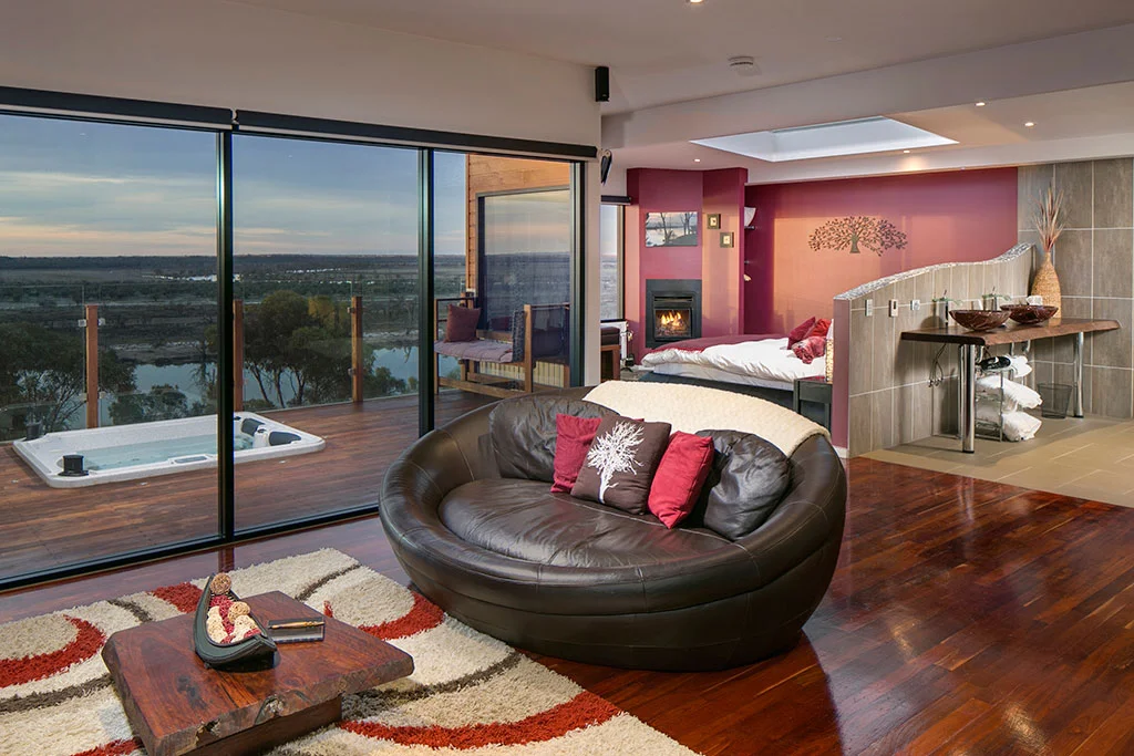 Open-plan villa interior with spa deck access, featuring fireplace, round leather sofa, polished timber floors and floor-to-ceiling windows overlooking river landscape