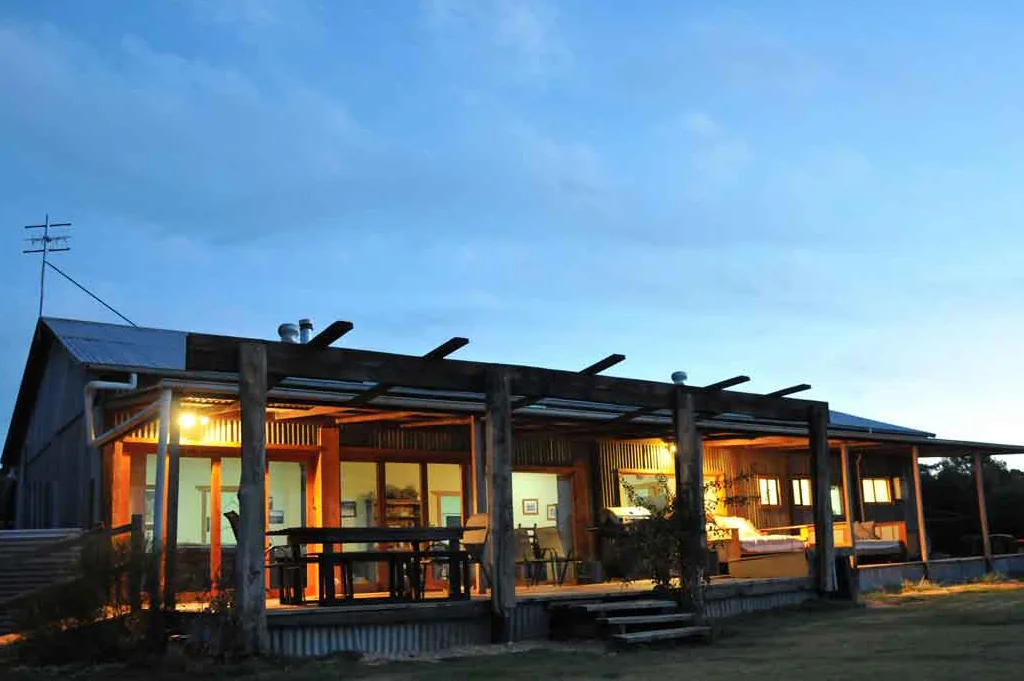 Illuminated sustainable luxury villa exterior at dusk with solar panels, modern pergola design and warm interior lighting in native Australian landscape
